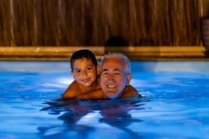 Grandson on grandfather's shoulder in pool at night